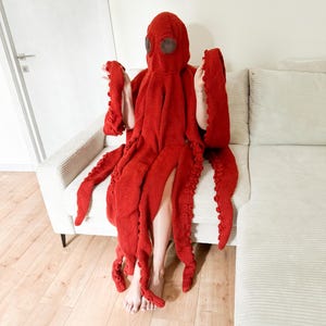 May include: A vibrant red octopus costume, featuring a hood with eye coverings and multiple tentacle-like appendages. The costume is made of a knitted material and is designed to be worn. The person is sitting on a light-colored couch.