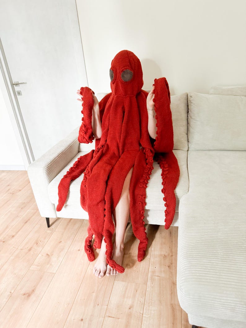 May include: A vibrant red octopus costume, featuring a hood with eye coverings and multiple tentacle-like appendages. The costume is made of a knitted material and is designed to be worn. The person is sitting on a light-colored couch.