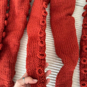 May include: Close-up of several rust-colored knitted tentacles, each with textured, circular details along the edges. The tentacles are arranged in a wavy pattern, showcasing the detailed knitwork and the rich, warm color.