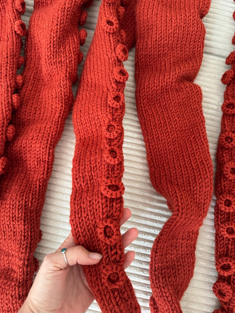 May include: Close-up of several rust-colored knitted tentacles, each with textured, circular details along the edges. The tentacles are arranged in a wavy pattern, showcasing the detailed knitwork and the rich, warm color.