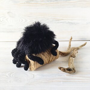 May include: A black crocheted spider toy sitting on a ball of brown yarn. The spider has a fuzzy body and long, spindly legs.