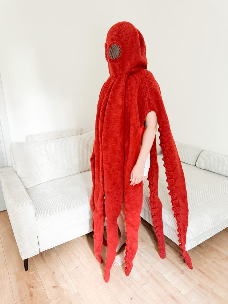May include: A vibrant red octopus costume with a hooded headpiece and long, tentacle-like appendages. The costume features a circular eye detail on the hood and is made of a knitted material. The tentacles extend to the floor, creating a unique and eye-catching design.