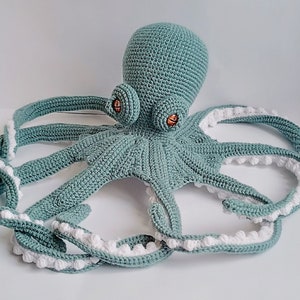 May include: A crocheted octopus plush toy with a teal blue body and white accents. The octopus has eight tentacles with white pom-poms along the edges.