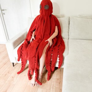 May include: A red knitted octopus costume with a hood and tentacles. The costume is worn by a person sitting on a light-colored couch. The tentacles are long and detailed.