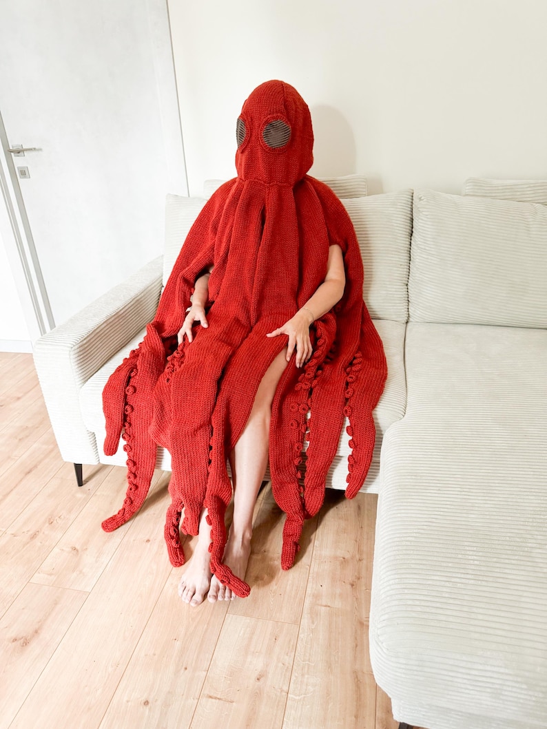 May include: A red knitted octopus costume with a hood and tentacles. The costume is worn by a person sitting on a light-colored couch. The tentacles are long and detailed.