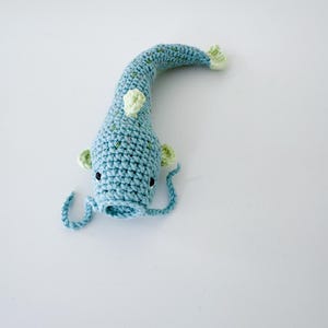 以下が含まれることがあります： 青と緑の色合いの手作りのかぎ針編みのナマズ。魚は、小さな緑色のアクセントと黒い目を持つ、長く湾曲した体を持っています。2本の長い青いひげがあり、柔らかい糸で作られています。