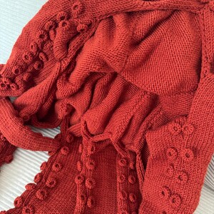 May include: A rust-colored knitted garment with a textured, organic design. The garment features a hood and sleeves, with decorative circular embellishments along the edges, creating a unique, tactile appearance. The knitwear is made of a soft, warm material.