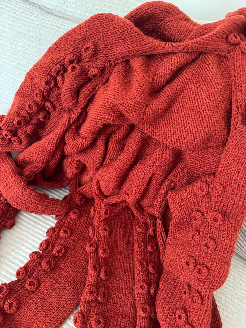 May include: A rust-colored knitted garment with a textured, organic design. The garment features a hood and sleeves, with decorative circular embellishments along the edges, creating a unique, tactile appearance. The knitwear is made of a soft, warm material.