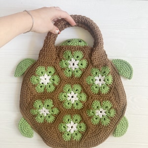 Puede incluir: Una bolsa de ganchillo de color marrón con forma de tortuga con detalles verdes y flores blancas. La bolsa tiene un asa y está hecha con un patrón de granny square.