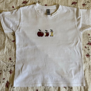 May include: White t-shirt with a red and yellow embroidered design of three apples, two whole and one with a bite taken out.