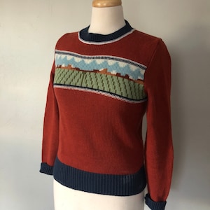 Peut inclure: Un pull couleur rouille avec un motif géométrique bleu, blanc et vert autour de l'encolure. Le pull a une bordure côtelée bleu marine autour de l'encolure et des poignets.