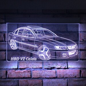 May include: An illuminated sign showing a white outline of a sedan car, with a sunroof and custom wheels. The text "HBD VZ Calais" is at the bottom. The sign is mounted on a brick wall. The car's design is highlighted.