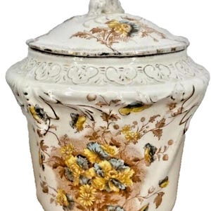 Pode incluir: Pote de cerâmica branco com tampa, com um padrão floral em amarelo, azul e castanho. O pote tem uma borda esculpida e uma pequena figura na tampa. O design é vintage e decorativo.