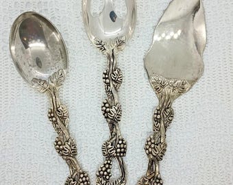 May include: Set of three silver-toned serving utensils. The spoon, fork, and cake server feature handles adorned with grape vine and leaf designs. The spoon has a rounded bowl, the fork has three tines, and the server has a wide, flat blade.