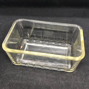 May include: A rectangular, clear glass dish with slightly rounded corners. The dish has a raised, decorative pattern along the bottom edge and a subtle yellow tint. The glass appears to be thick and sturdy, suitable for baking or serving.
