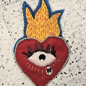 May include: A red felt heart with a white eye and a single teardrop, surrounded by black eyelashes and beads. The heart is topped with a yellow flame with blue edges.