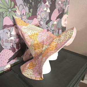 May include: A pointed hat featuring a floral pattern in shades of pink, orange, yellow, and green. The fabric has a dense print of small flowers covering the entire surface. The text "Stitchanddie_creations" is printed on the side of the hat.