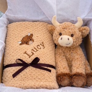 May include: A tan-colored baby blanket with the name "Lucy" embroidered on it, along with a brown Highland cow graphic. A matching plush Highland cow toy sits beside the blanket in a gift box, tied with a brown ribbon.