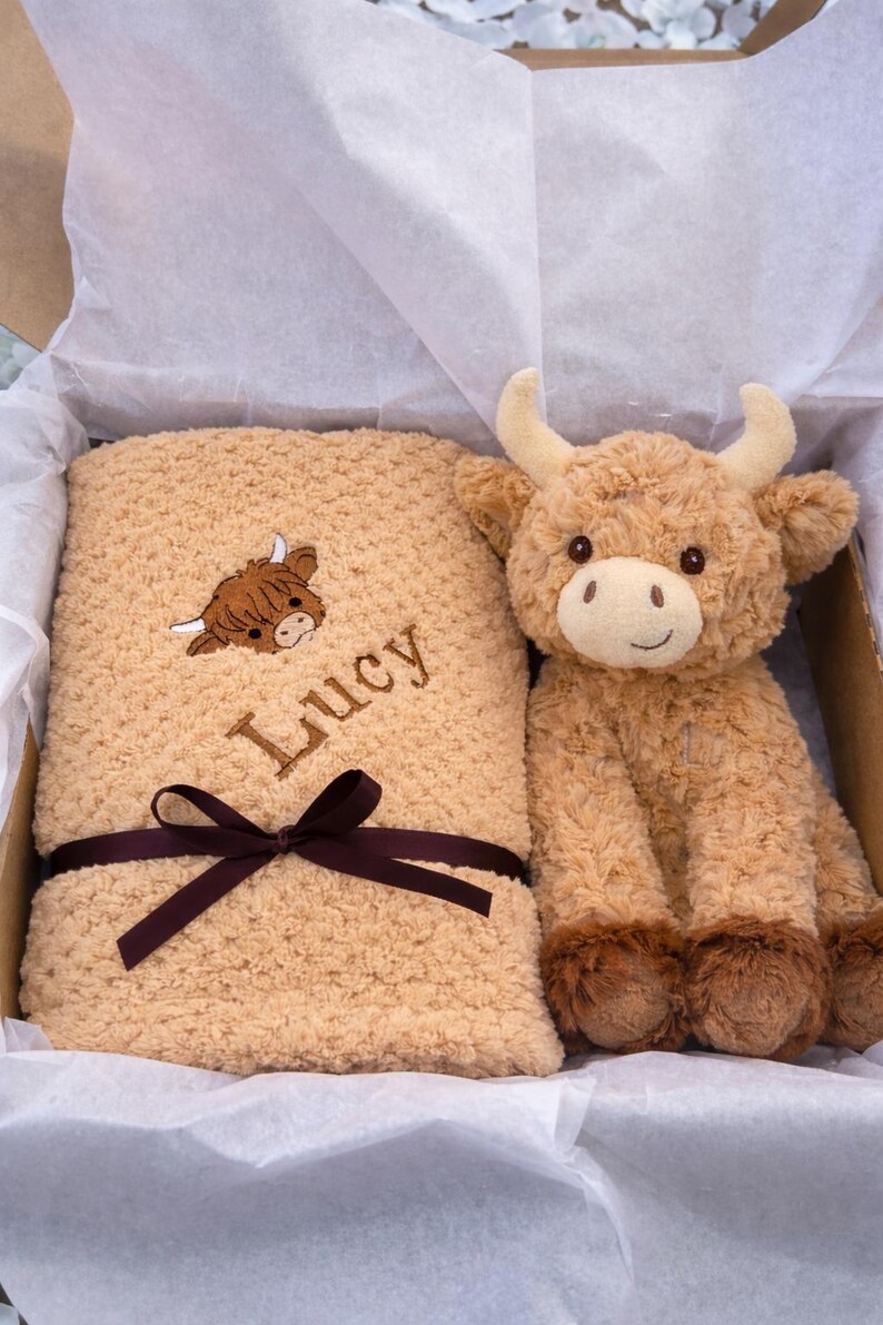 May include: A tan-colored baby blanket with the name "Lucy" embroidered on it, along with a brown Highland cow graphic. A matching plush Highland cow toy sits beside the blanket in a gift box, tied with a brown ribbon.