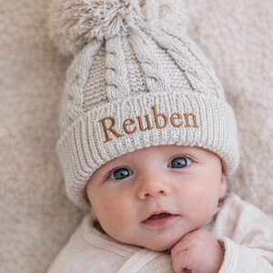 May include: A beige cable-knit beanie with a large matching pom-pom on top. The cuff of the hat features the name Reuben embroidered in brown thread. The hat has a textured knit pattern and a folded brim, providing a warm and cosy appearance.