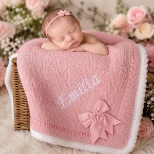 May include: A sleeping baby wearing a pink headband rests on a pink cable-knit blanket. The blanket has the name "Emilia" embroidered in white and a pink bow. The blanket is in a wicker basket.