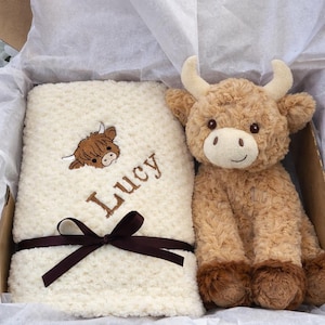 May include: A gift set featuring a plush Highland cow and a cream-colored blanket. The blanket is embroidered with a Highland cow graphic and the name "Lucy" in brown thread. A brown ribbon is tied around the blanket. The cow plush is brown and tan.