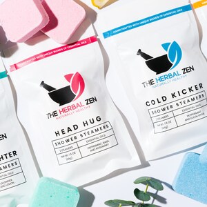 May include: Four white pouches with different essential oil shower steamers. Each pouch features a black mortar and pestle logo with a green leaf and the text "The Herbal Zen Naturally Healthy". The pouches are labeled with different scents and names: "Stress Fighter", "Head Hug", "Cold Kicker", and "Tropical". The pouches are surrounded by eucalyptus leaves and blue and pink bath bombs.
