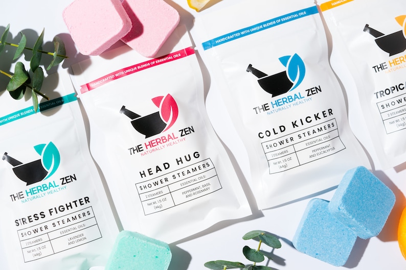 May include: Four white pouches with different essential oil shower steamers. Each pouch features a black mortar and pestle logo with a green leaf and the text "The Herbal Zen Naturally Healthy". The pouches are labeled with different scents and names: "Stress Fighter", "Head Hug", "Cold Kicker", and "Tropical". The pouches are surrounded by eucalyptus leaves and blue and pink bath bombs.