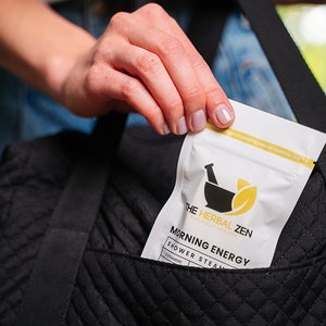 May include: A white pouch with the text "The Herbal Zen" and "Morning Energy Shower Steam" in black text. The pouch is being placed into a black quilted bag.