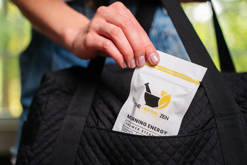 May include: A white pouch with the text "The Herbal Zen" and "Morning Energy Shower Steam" in black text. The pouch is being placed into a black quilted bag.