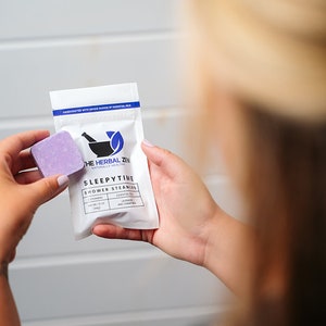 May include: A white pouch with a purple shower steamer inside. The pouch has the text "The Herbal Zen" and "Sleepytime Shower Steamers" printed on it. The shower steamer is lavender and chamomile scented.