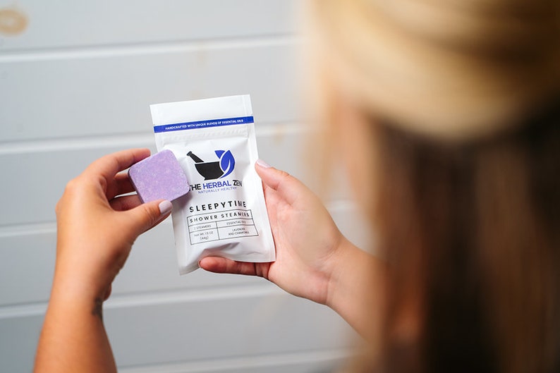 May include: A white pouch with a purple shower steamer inside. The pouch has the text "The Herbal Zen" and "Sleepytime Shower Steamers" printed on it. The shower steamer is lavender and chamomile scented.