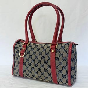 May include: A Gucci handbag featuring a beige canvas body with a repeating dark blue GG monogram pattern. The bag includes red leather trim, two red leather top handles, gold-tone hardware, and a gold-tone D-ring detail on the front panel.