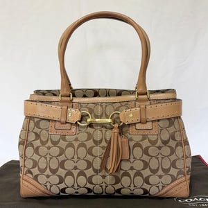 Op de afbeelding: Een beige en bruine handtas met monogramprint, een gestructureerd silhouet, twee leren handvatten en een horizontale leren band aan de voorzijde met goudkleurige metalen details. Een leren kwastje hangt aan de centrale gesp.