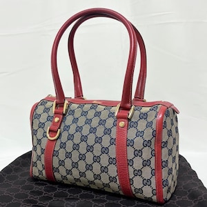 May include: A cylindrical handbag with red leather handles and trim. The bag features a beige and navy blue pattern with gold-colored hardware. The bag is sitting on a black surface.