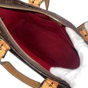 Może przedstawiać: Brązowa torebka z monogramem, z czerwonym wnętrzem. Torba posiada zapięcie na zamek błyskawiczny oraz brązowe sk&oacute;rzane uchwyty i wykończenia. Wnętrze jest widoczne, pokazując kieszenie i przestronną komorę.