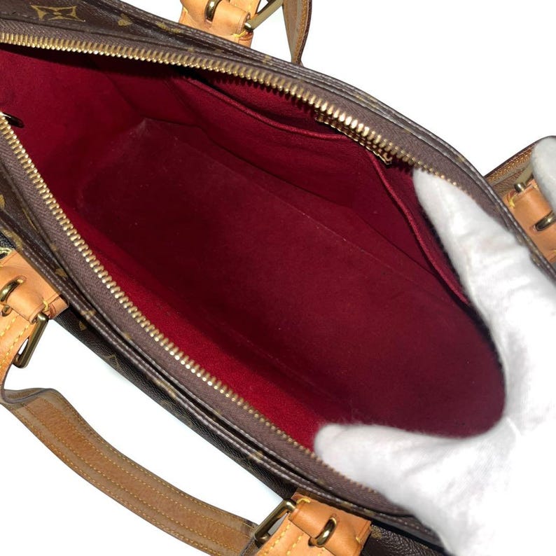 Może przedstawiać: Brązowa torebka z monogramem, z czerwonym wnętrzem. Torba posiada zapięcie na zamek błyskawiczny oraz brązowe sk&oacute;rzane uchwyty i wykończenia. Wnętrze jest widoczne, pokazując kieszenie i przestronną komorę.