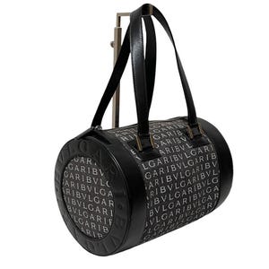 May include: Black and white cylindrical handbag with the brand name "BVLGARI" printed repeatedly. The bag has black leather handles and trim, with gold-colored metal accents. The bag is displayed on a silver metal stand.