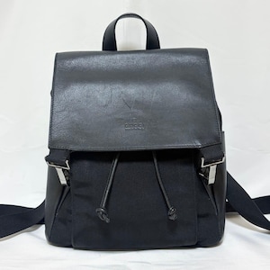 Puede incluir: Una mochila negra con una solapa superior de cuero liso que presenta el texto en relieve GUCCI made in italy. El cuerpo principal está fabricado en tela negra con cierre de cordón, dos correas negras para los hombros y un asa superior.