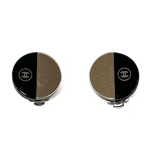 May include: A pair of round clip-on earrings. Each earring is divided in half, with one side black and the other silver. The black side features a white interlocking CC logo. The earrings have a silver clip-on backing.