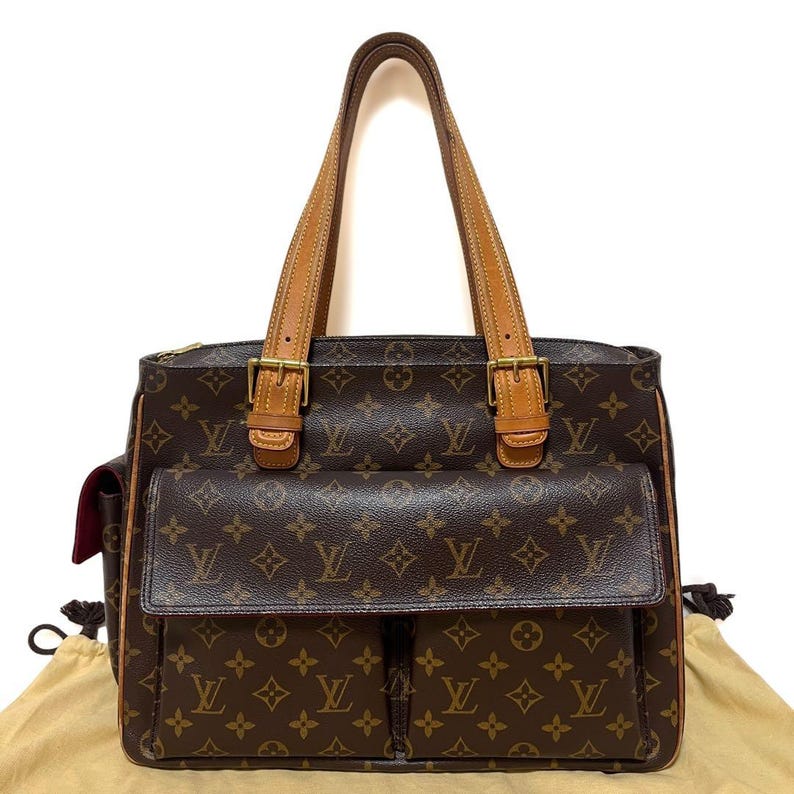 Może przedstawiać: Brązowa torba typu tote z monogramem z pł&oacute;tna z dwiema przednimi kieszeniami i jasnobrązowymi sk&oacute;rzanymi akcentami. Torba posiada dwa g&oacute;rne uchwyty, złote okucia i zapięcie na zamek błyskawiczny. Wnętrze jest jasnobrązowe.