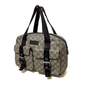 May include: A beige and brown Gucci handbag with the iconic GG monogram pattern. The bag features two front pockets with silver buckles, black straps, and a top handle. The bag is designed with a rectangular shape and a zipper closure.