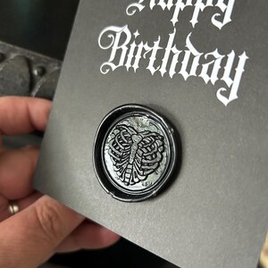 May include: A dark gray birthday card with the words "Happy Birthday" in white gothic script. A black wax seal with a detailed rib cage design is affixed to the card, adding a unique touch.