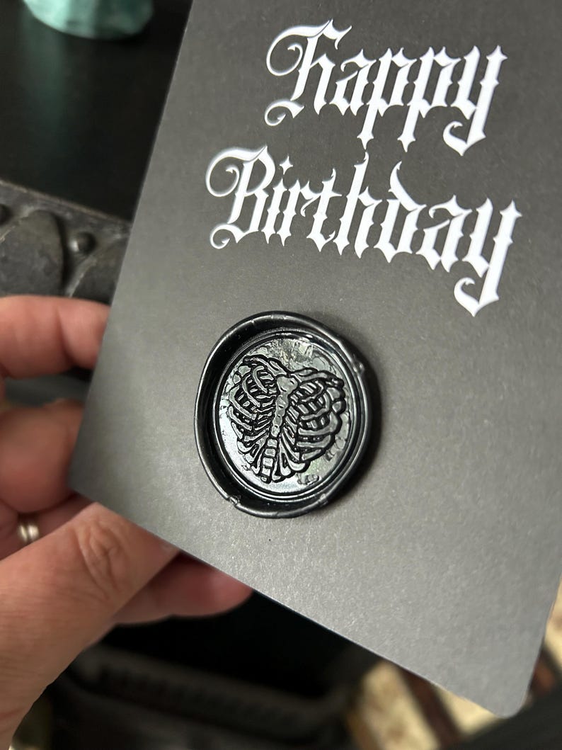 May include: A dark gray birthday card with the words "Happy Birthday" in white gothic script. A black wax seal with a detailed rib cage design is affixed to the card, adding a unique touch.