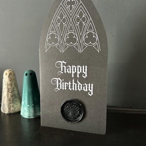 May include: A dark gray birthday card with a gothic arch shape, featuring white ornate designs at the top and the words "Happy Birthday." A black wax seal is affixed at the bottom. Two small ceramic ghost figurines are in the background.