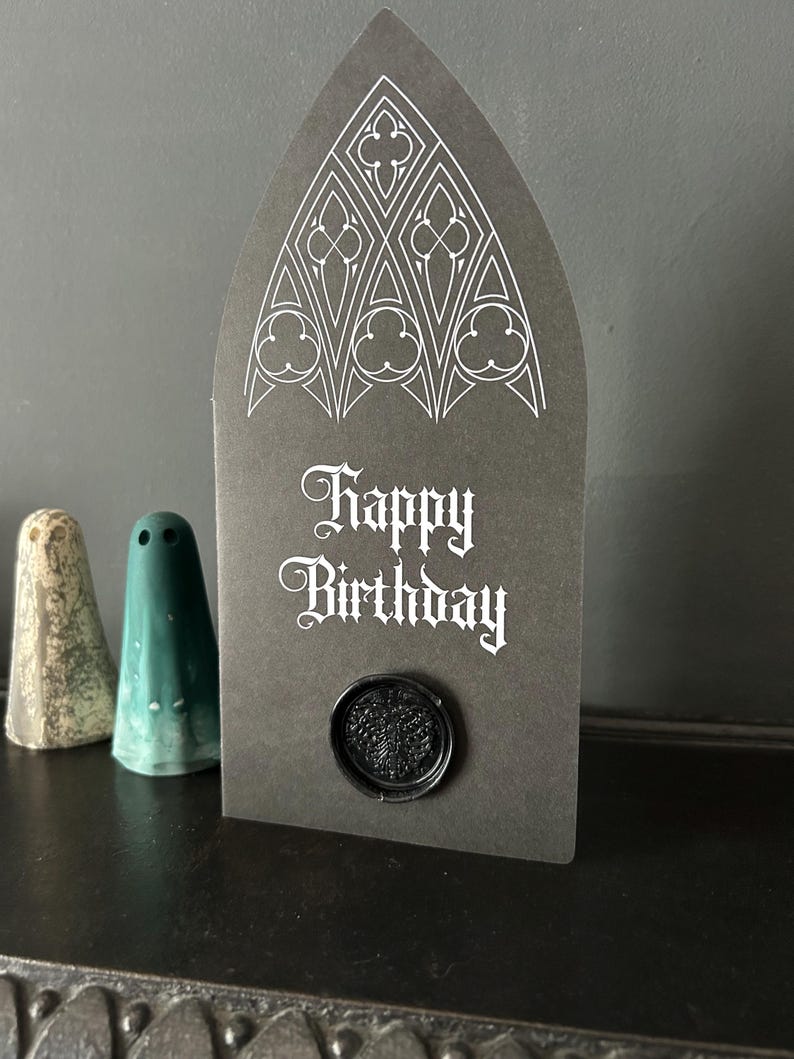 May include: A dark gray birthday card with a gothic arch shape, featuring white ornate designs at the top and the words "Happy Birthday." A black wax seal is affixed at the bottom. Two small ceramic ghost figurines are in the background.