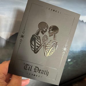 May include: A dark grey card with a silver embossed design of two skeletons holding hands. The text "'Til Death" is below the skeletons. The card is decorated with crescent moons and stars.