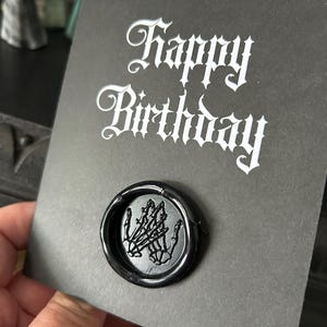 May include: A black greeting card with the words "Happy Birthday" in white gothic script. A black wax seal with a skeleton hand design is affixed to the card. The card is held in a hand.