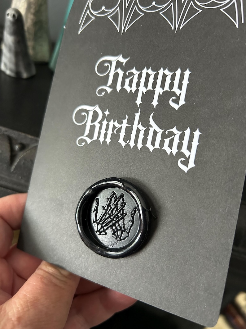 May include: A black greeting card with the words "Happy Birthday" in white gothic script. A black wax seal with a skeleton hand design is affixed to the card. The card is held in a hand.