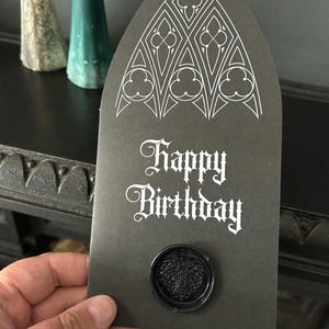 May include: A black birthday card with a gothic arch design and the words "Happy Birthday" in white script. The card has a black wax seal with a leaf design. Two ghost-shaped figurines are in the background.